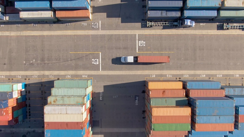White truck transports a red freight container across the busy port of Los Angeles. Flying above a freight truck hauling a heavy shipment past large stacks of other containers. 