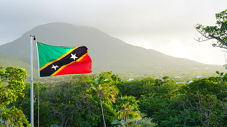 An image of the flag of the St Kitts and Nevis floating in front of the Nevis volcano.