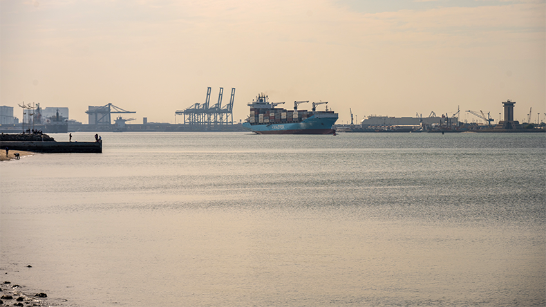 A Maersk cargoship sails steadily through the calm waters of Kuwait's harbour, heading toward a bustling port lined with towering cranes and industrial structures