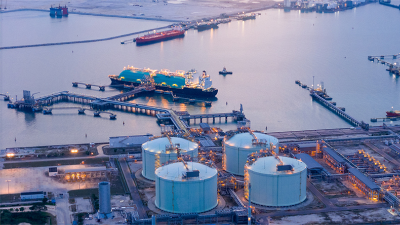 Aerial view of an LNG tanker docked at an offshore terminal with multiple pipelines, storage tanks, and an industrial complex. Captures global energy logistics